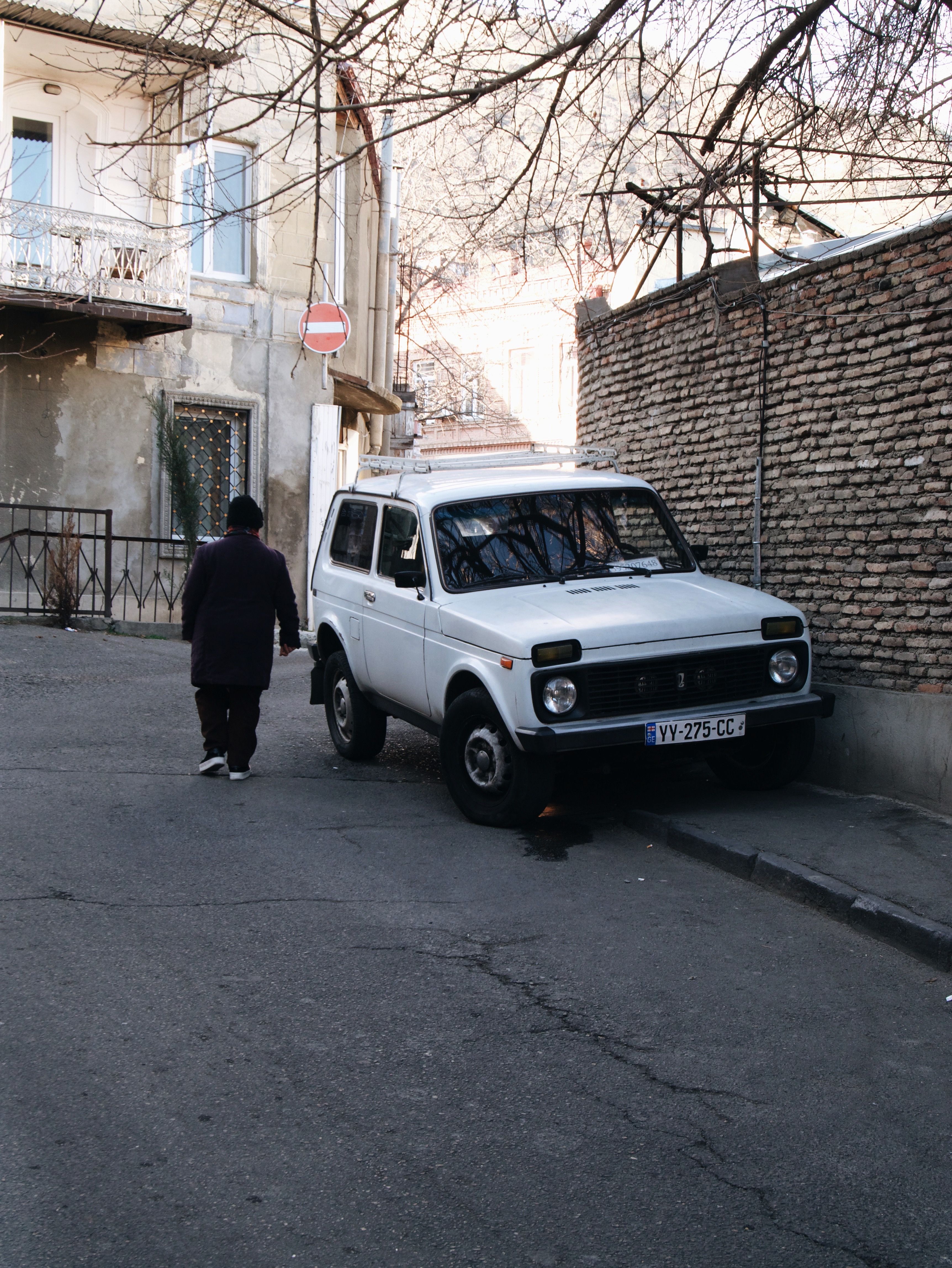 A vintage car parked on a winter street in Tbilisi, Georgia, with an adult walking nearby.