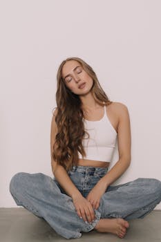 Young woman with wavy hair in white top and jeans sitting peacefully with eyes closed.