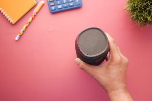 A hand holding a portable speaker on a colorful desk with a calculator, notebook, and plant.