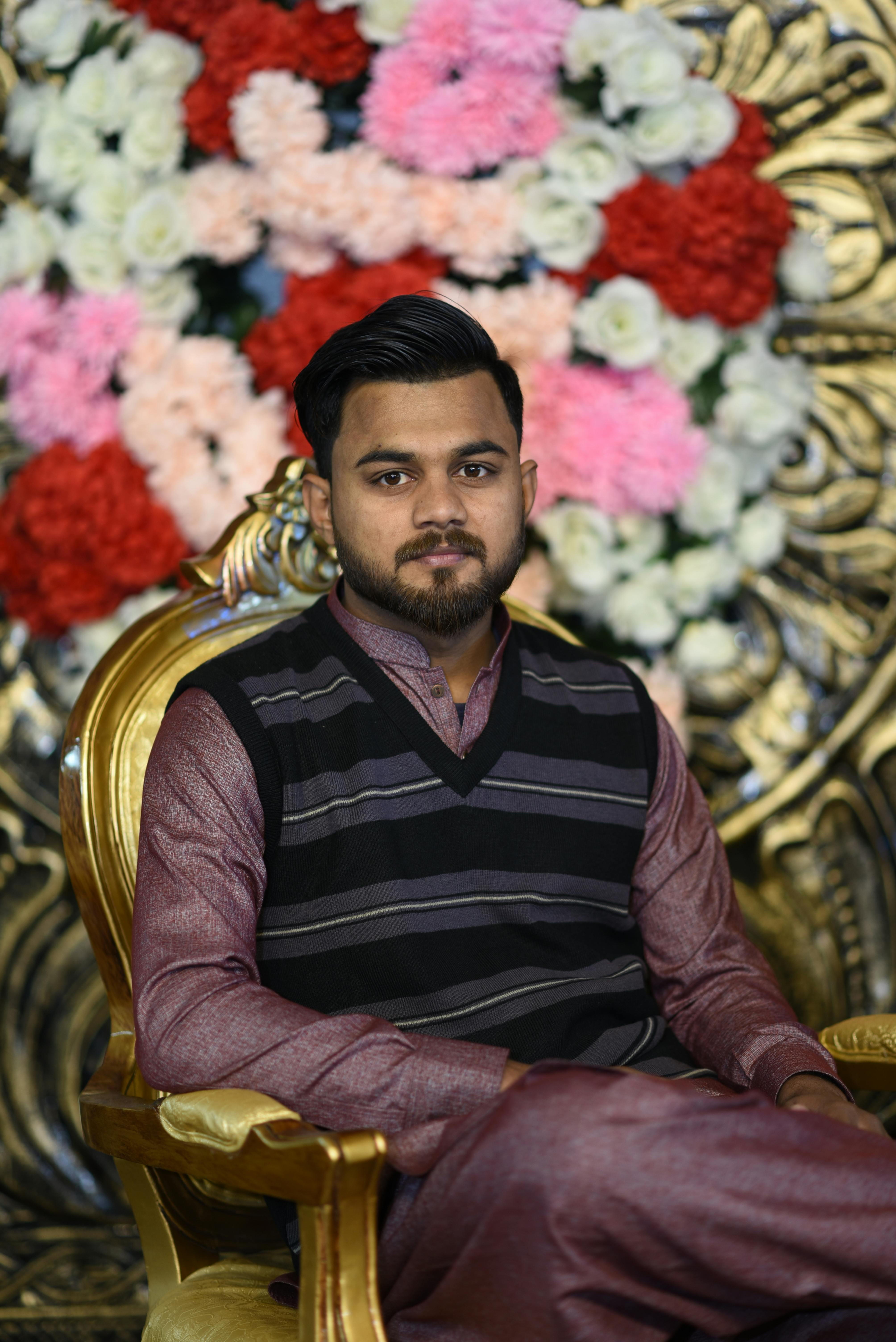 Man sitting on a golden armchair, in traditional attire, with a floral backdrop.