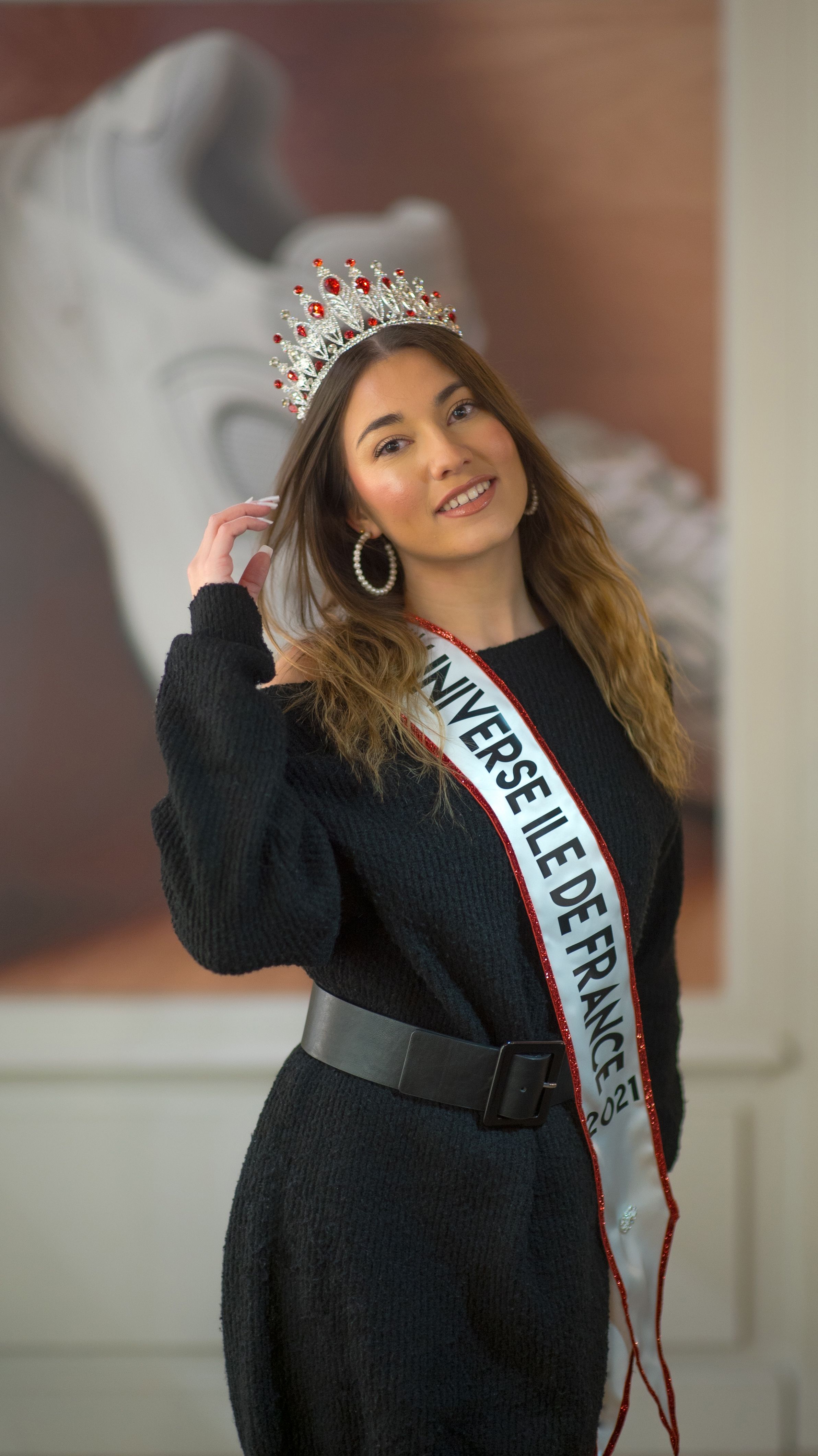 Woman smiling in a black dress and crown with a sash, posing indoors.