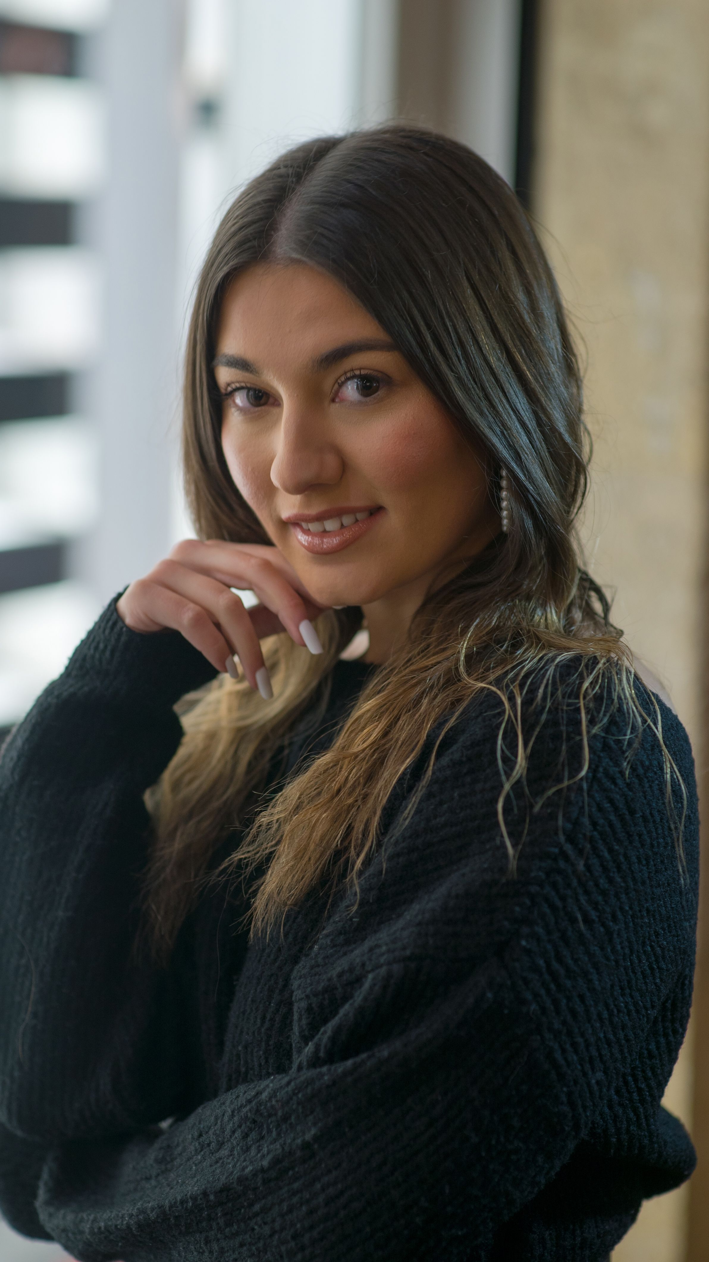 Close-up portrait of a smiling woman posing confidently, wearing a black sweater indoors.