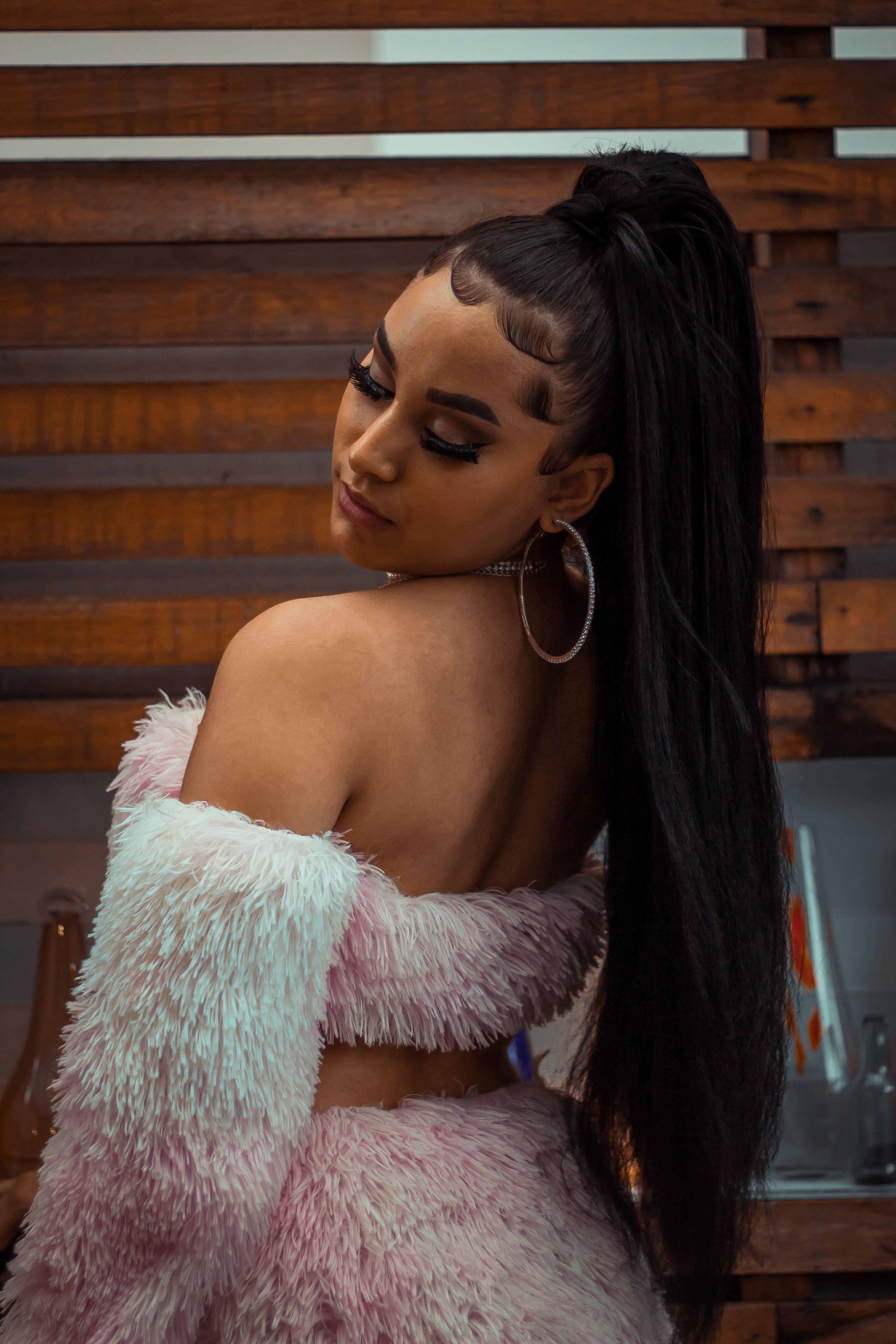 Elegant woman in pink fur outfit poses stylishly with long hair and hoop earrings, looking over her shoulder.