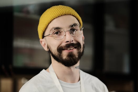 A bearded man wearing glasses and a yellow hat smiling indoors.