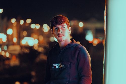 Free Young man in a casual hoodie poses at night with vibrant urban bokeh lights in the background. Stock Photo