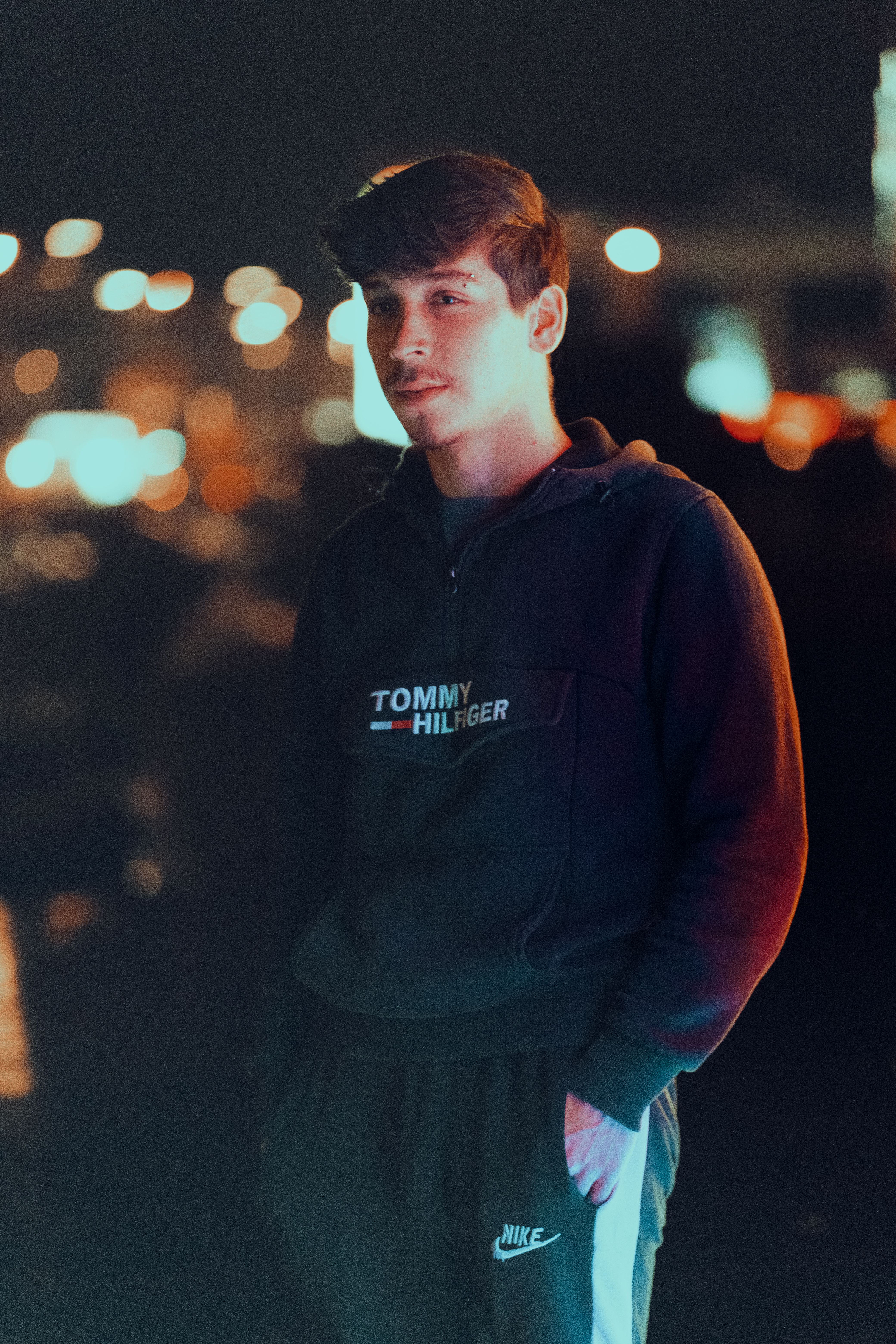 Portrait of a young man in a hoodie with city lights softly glowing in the background.