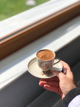 A hand holding a decorative porcelain cup of Turkish coffee near a sunlit window.