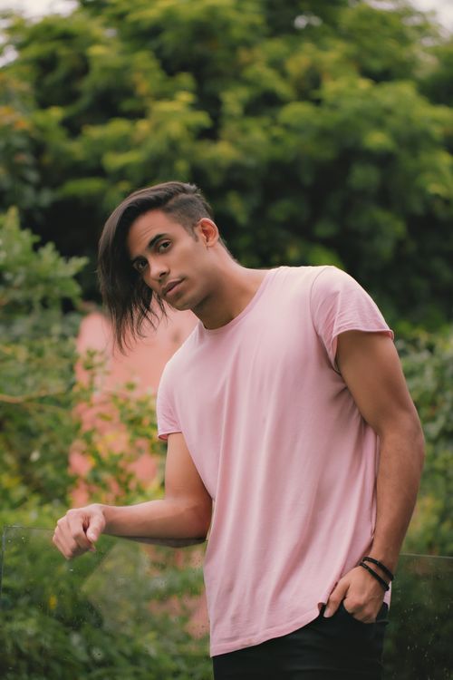 Free A young man in a pink t-shirt stands outdoors with a relaxed pose against a lush green background. Stock Photo