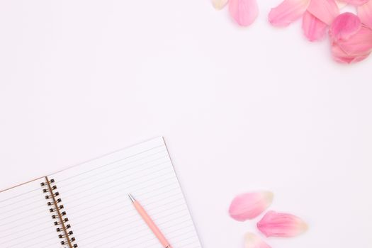 Flat lay of an open notebook with pink petals and a pen on a white background.