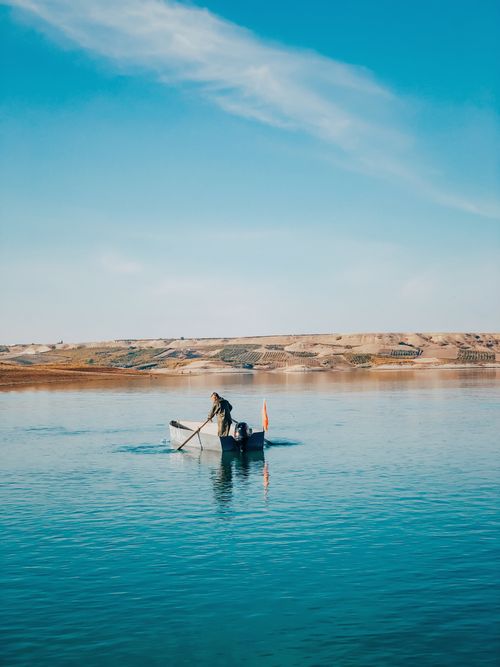 Free A lone fisherman paddles a small boat across a tranquil lake under clear blue skies. Stock Photo