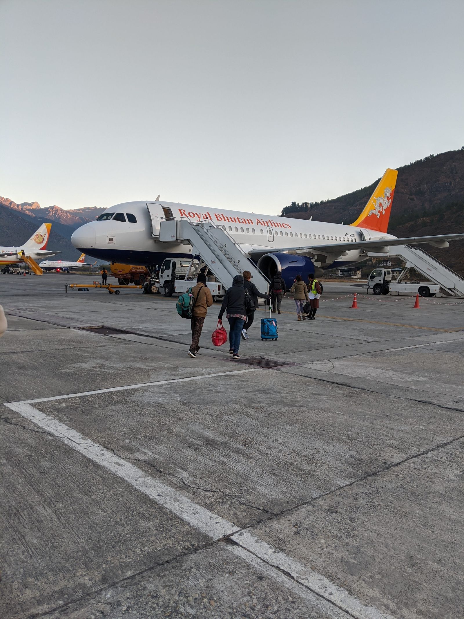 Travellers with bags crossing the Paro airport apron to board a Drukair aircraft, dry pine hills behind