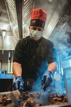 Chef wearing red hat and gloves cooking meat on a grill in a professional kitchen.