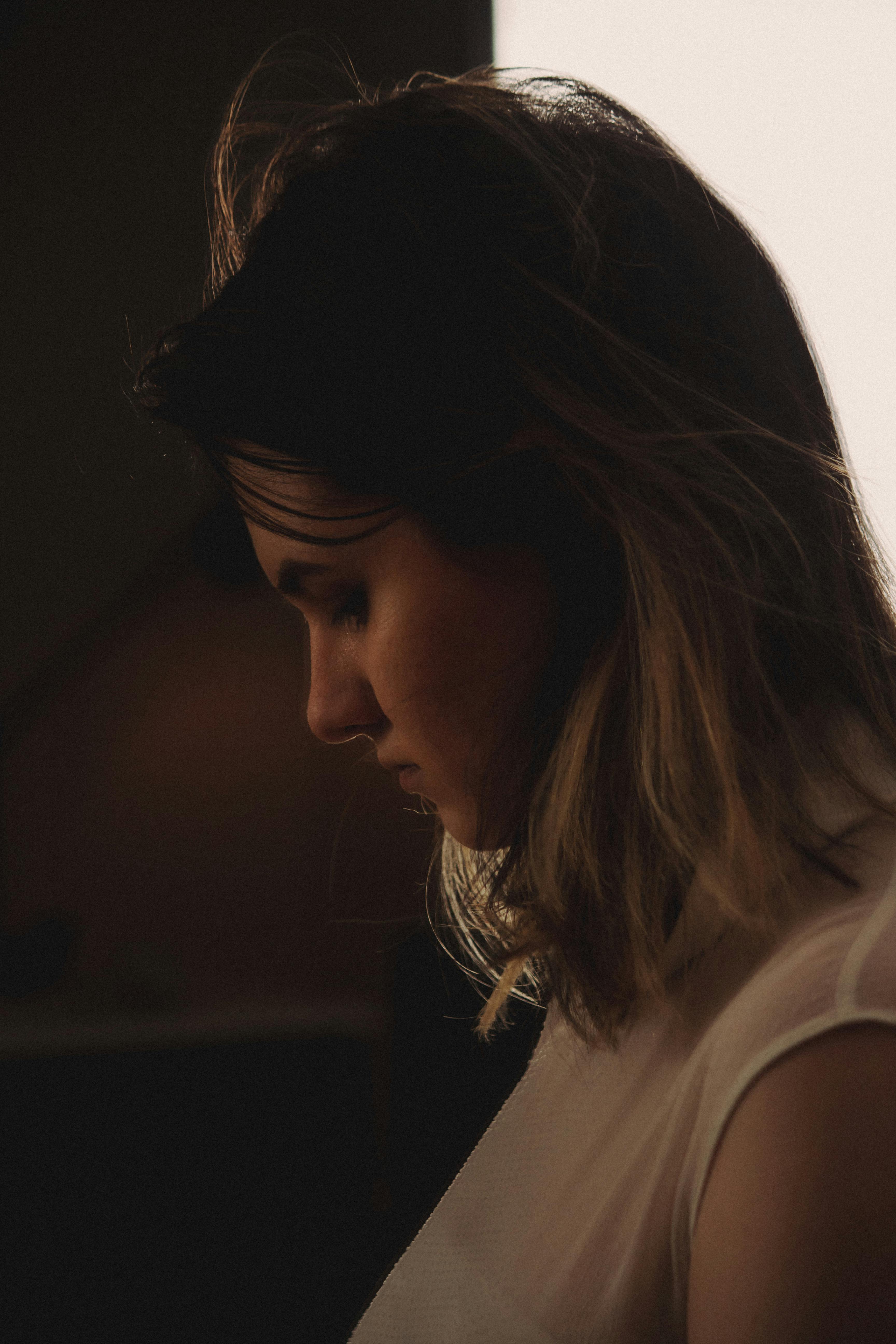 A moody, side profile portrait of a woman in a sleeveless white top captured indoors.