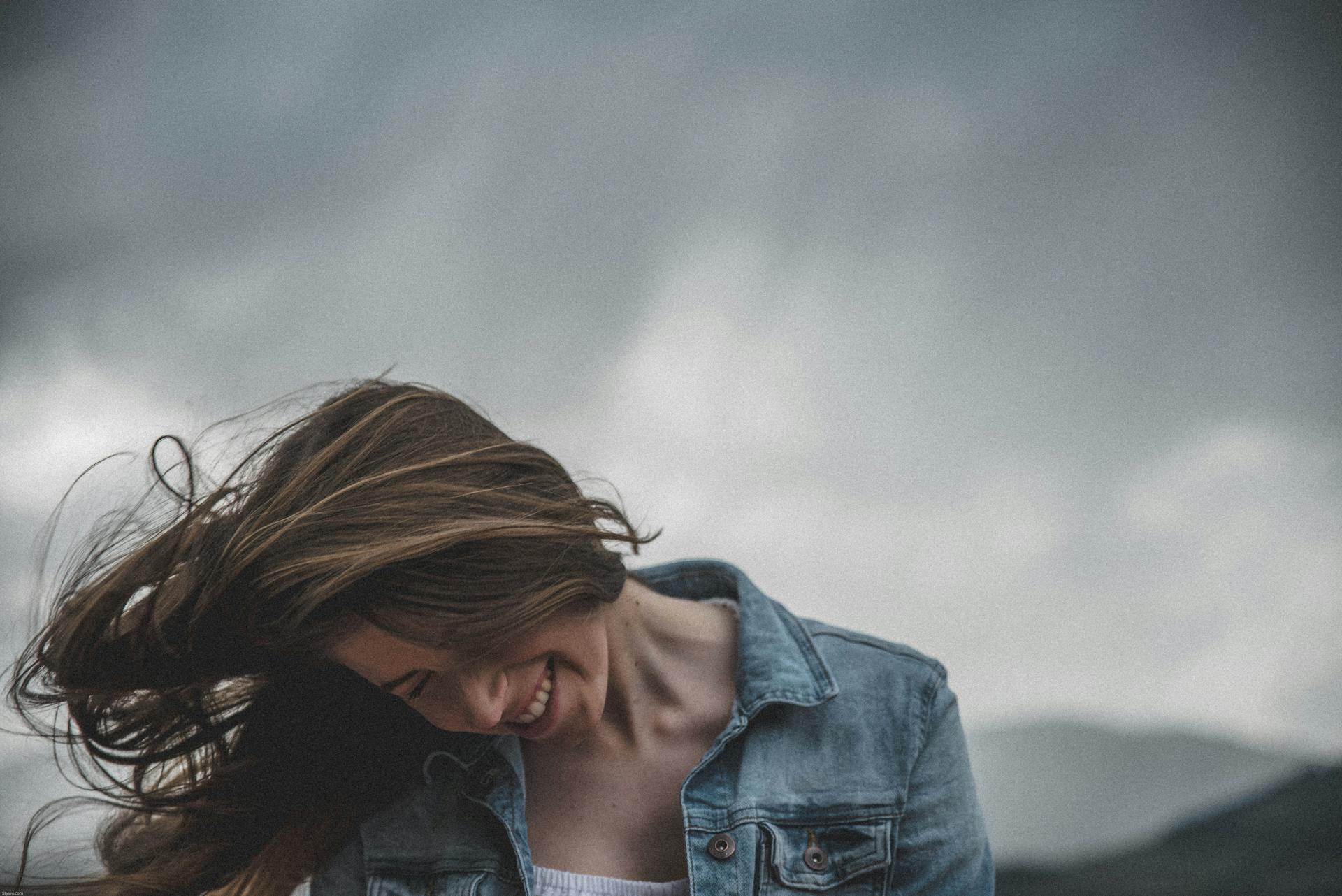 Young woman in denim jacket smiling and flipping hair in windy, cloudy weather in Kitzbühel.