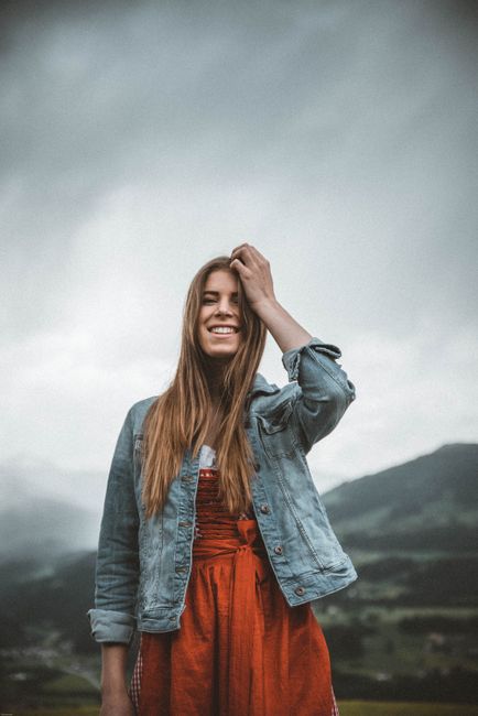 A young woman in a denim jacket smiling outdoors against a cloudy sky in Kitzbühel, Austria.