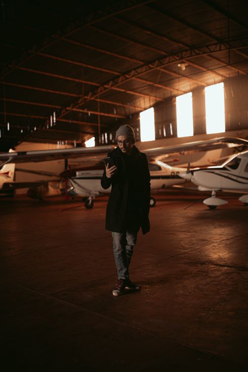 Free A man stands in an aircraft hangar, using his smartphone. Warm lighting highlights the planes. Stock Photo