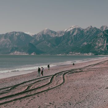 A tranquil beach scene with mountains, featuring people strolling and tire tracks on the sand.