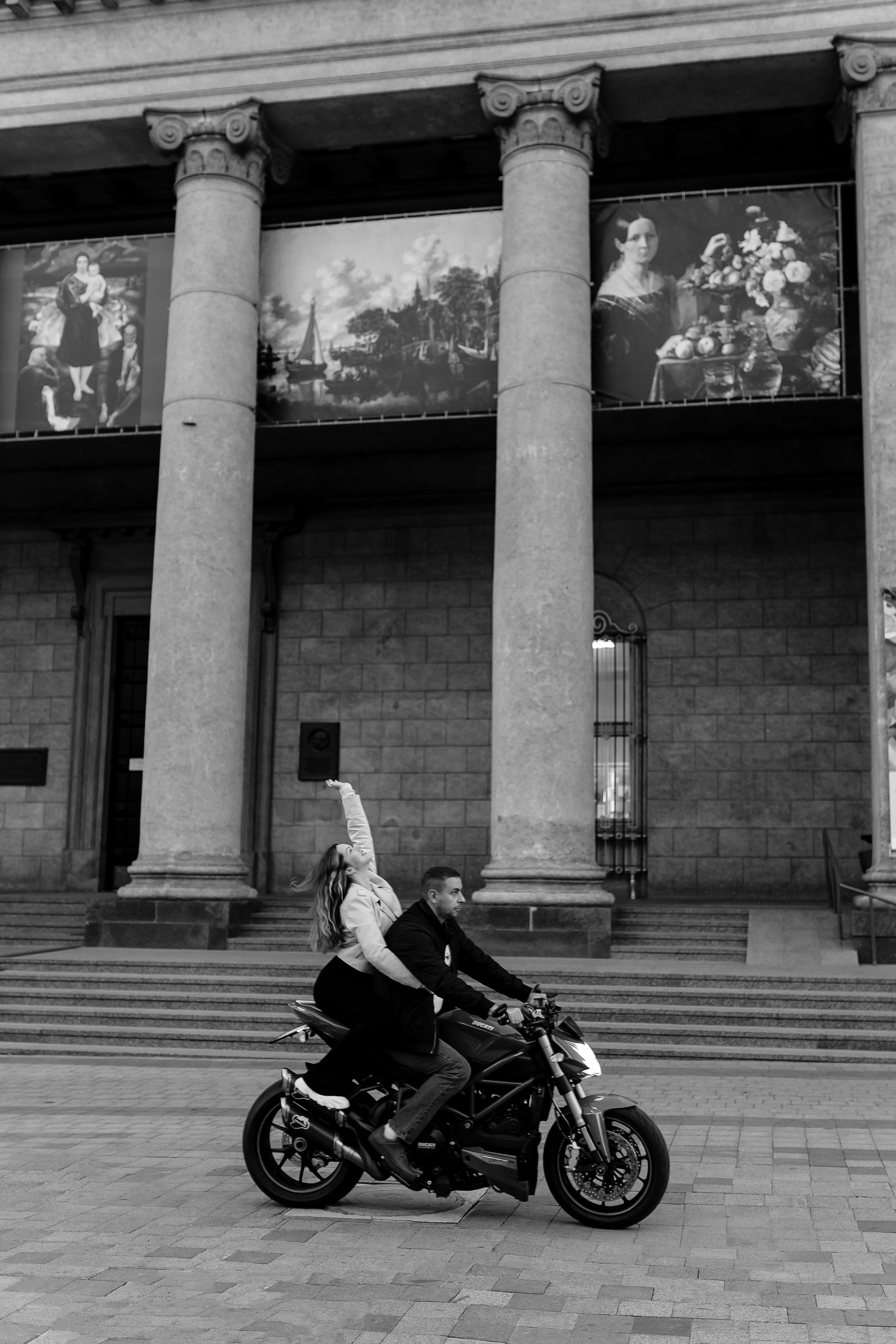 Monochrome image of a man and woman riding a motorcycle past a historic building with columns.