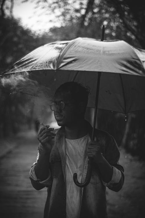 Grayscale Photo of a Man Smoking a Cigarette while Holding an Umbrella