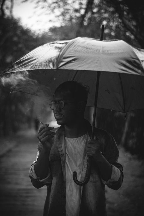Free A moody grayscale photo of a man smoking under an umbrella in the rain. Stock Photo