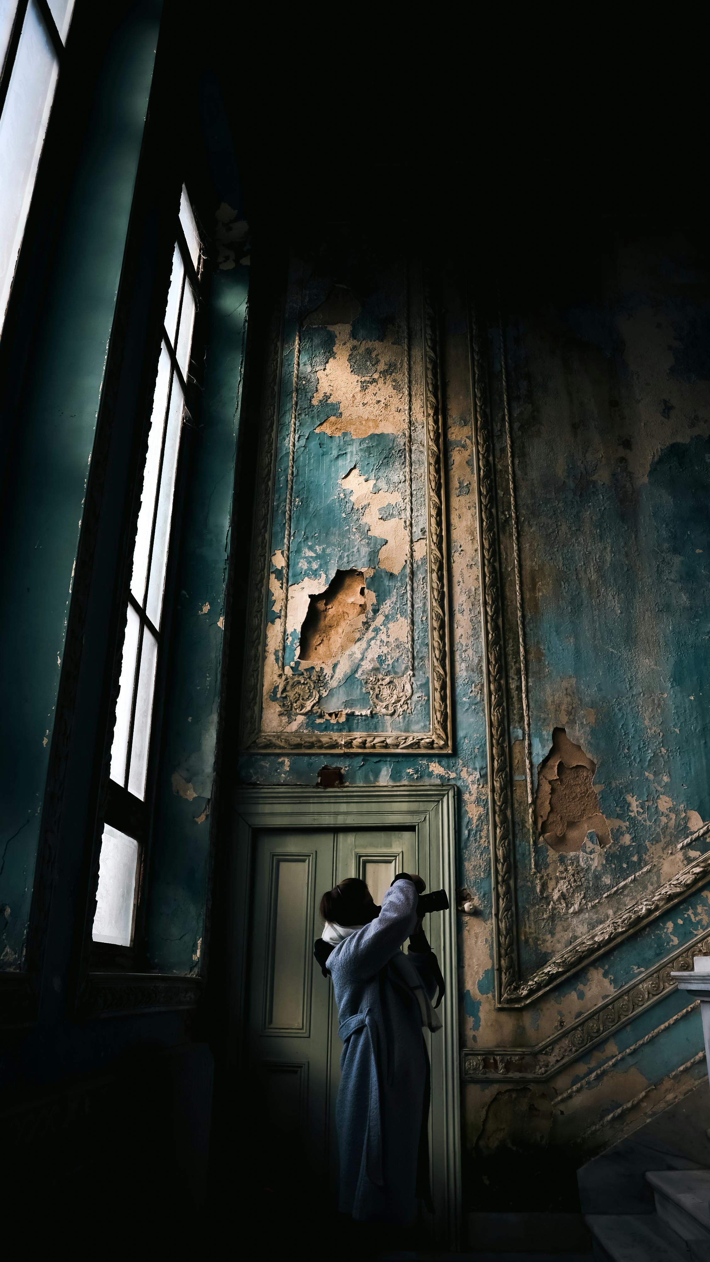 A person in coat stands in an abandoned mansion with chipped paint walls, creating a moody atmosphere.