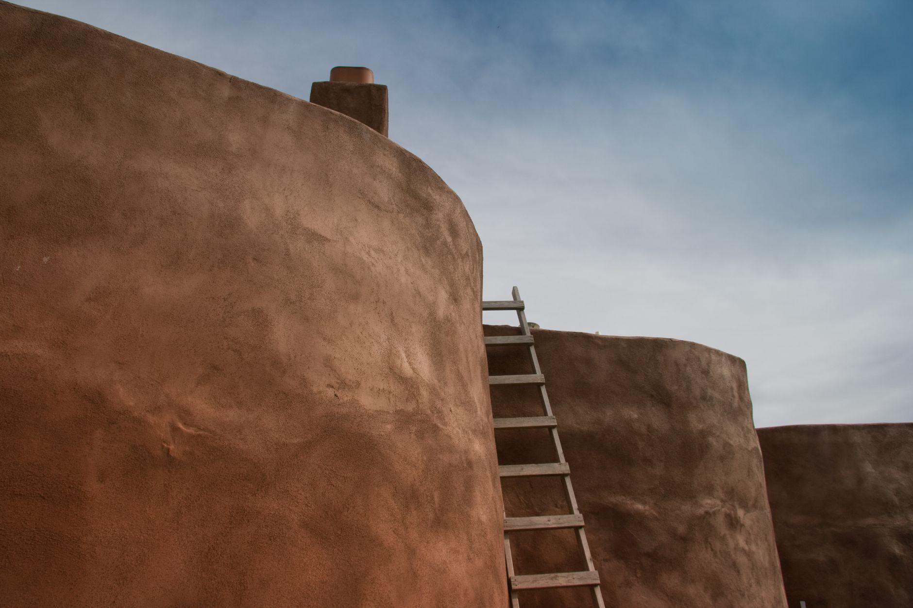 A rustic adobe building with a wooden ladder under a blue sky in New Mexico.