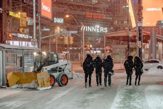 People walking on a snowy city street during a nighttime snowfall, with bright neon lights in the background.