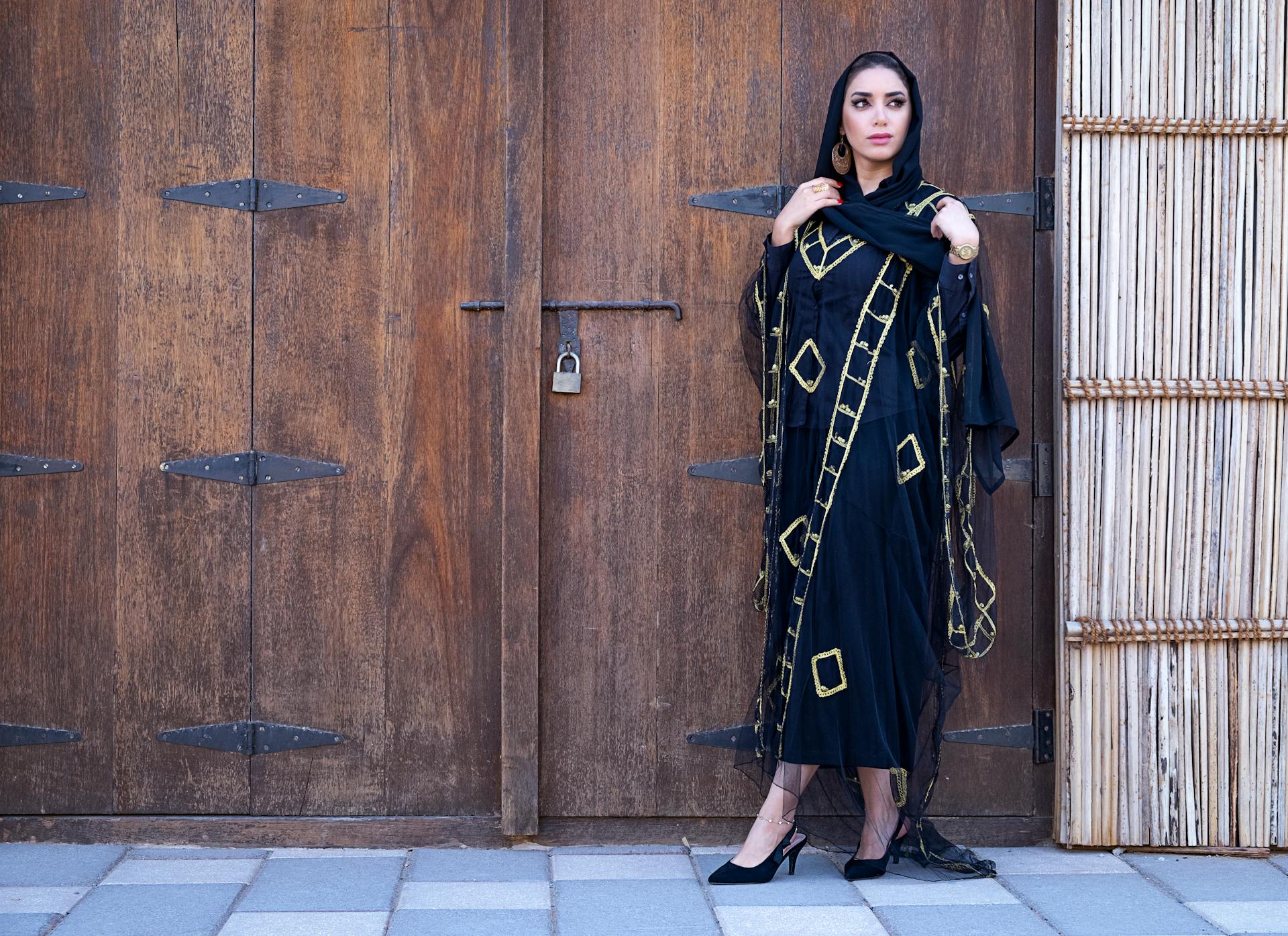 Middle Eastern woman in a black and gold traditional dress standing by a wooden door in Dubai.