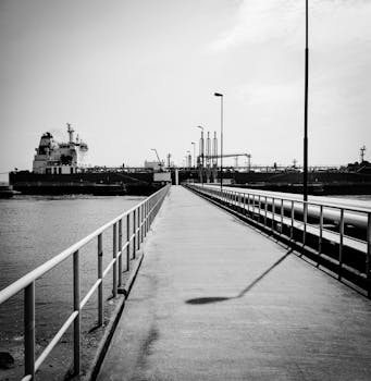 Black and white photo of a ship docked at an industrial harbor with a long concrete walkway.