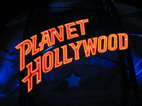 Illuminated Planet Hollywood neon sign against a dark blue background, featuring iconic vibrant lettering.