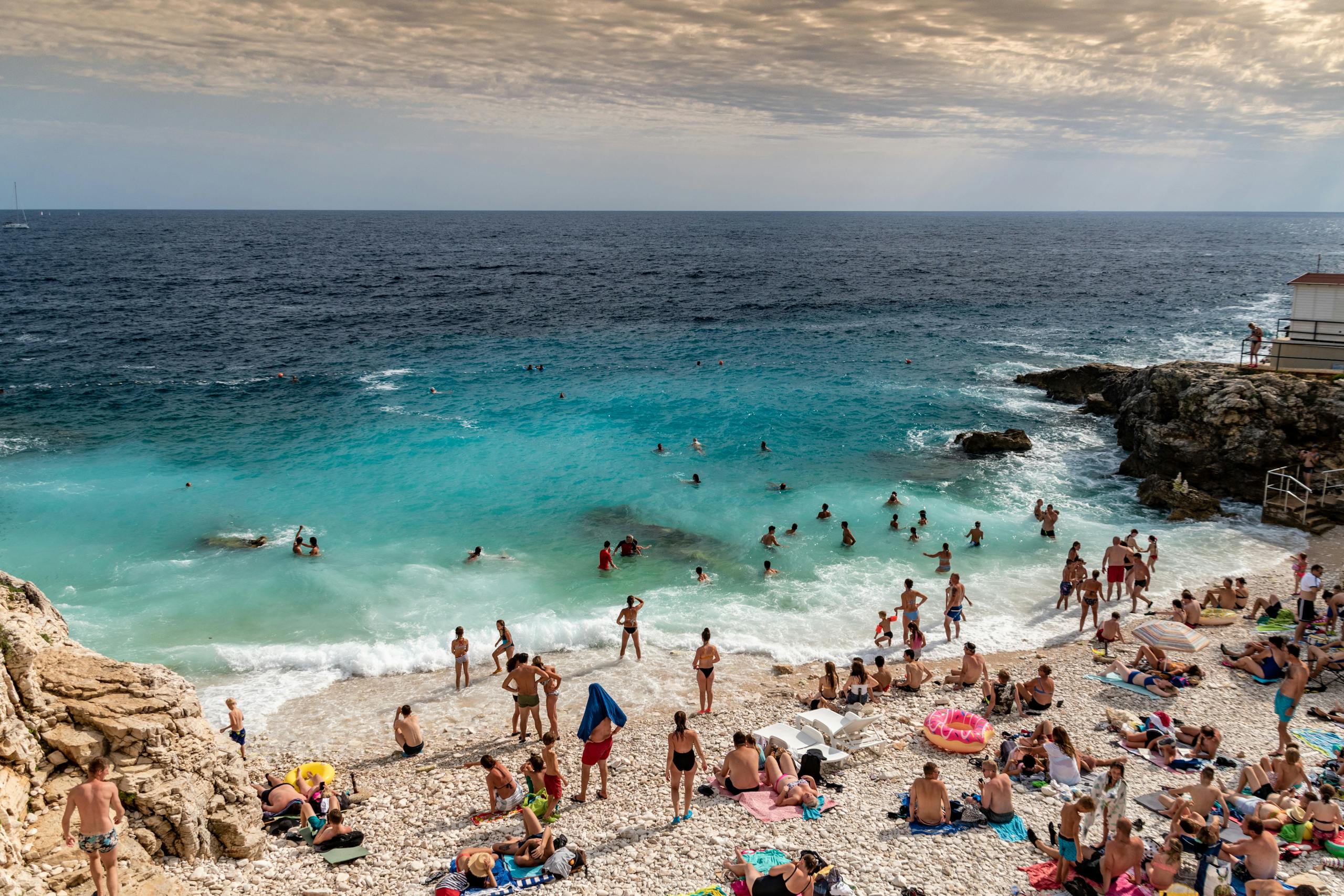 Sunbathers and swimmers enjoy a summer day at Pula beach, Croatia, with clear blue sea and rocky shore. by Sun Pixel Photography