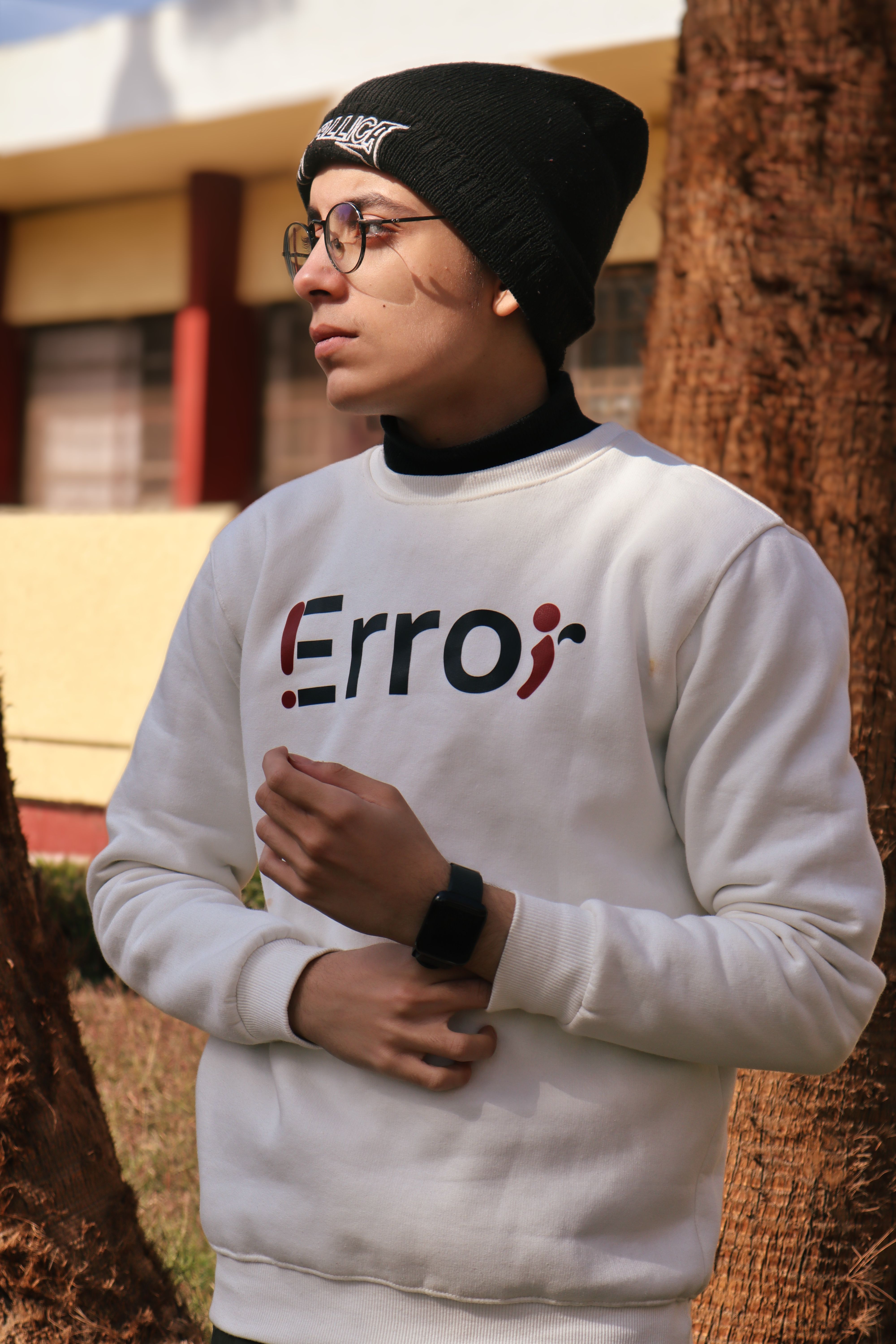 Young man wearing a beanie and sweater, posing outdoors. Trendy with eyeglasses and smartwatch.