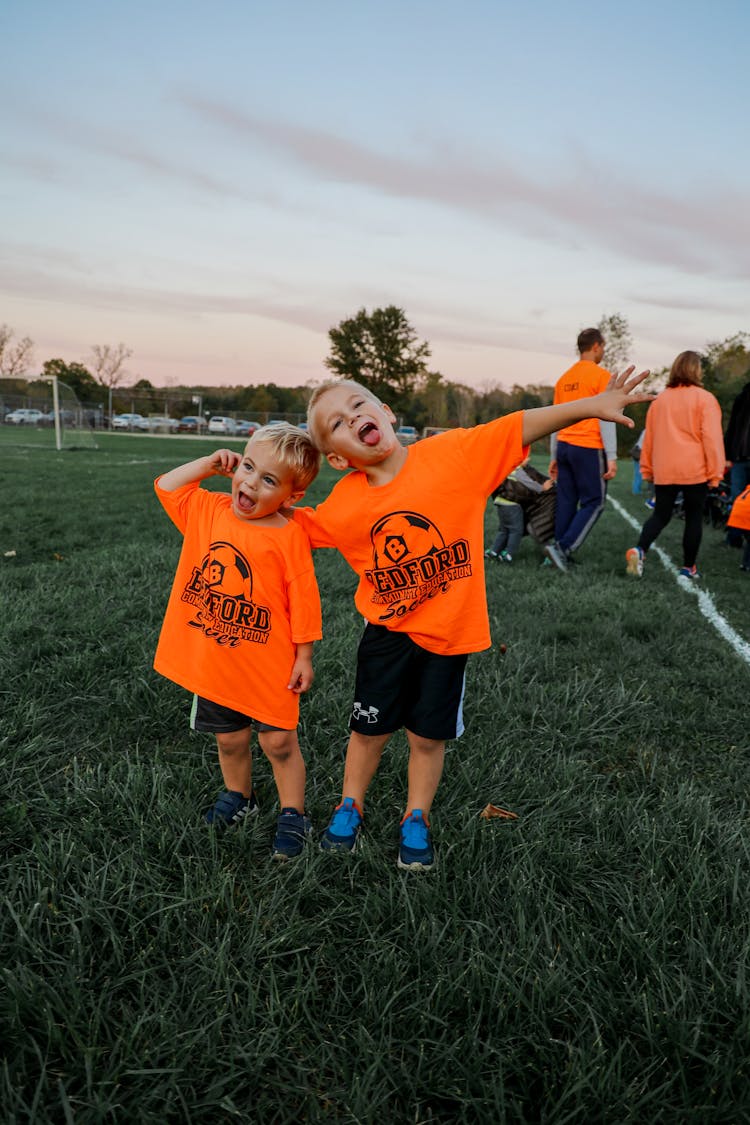 Two Adorable Boys In Orange Shirts Standing On Green Grass Field