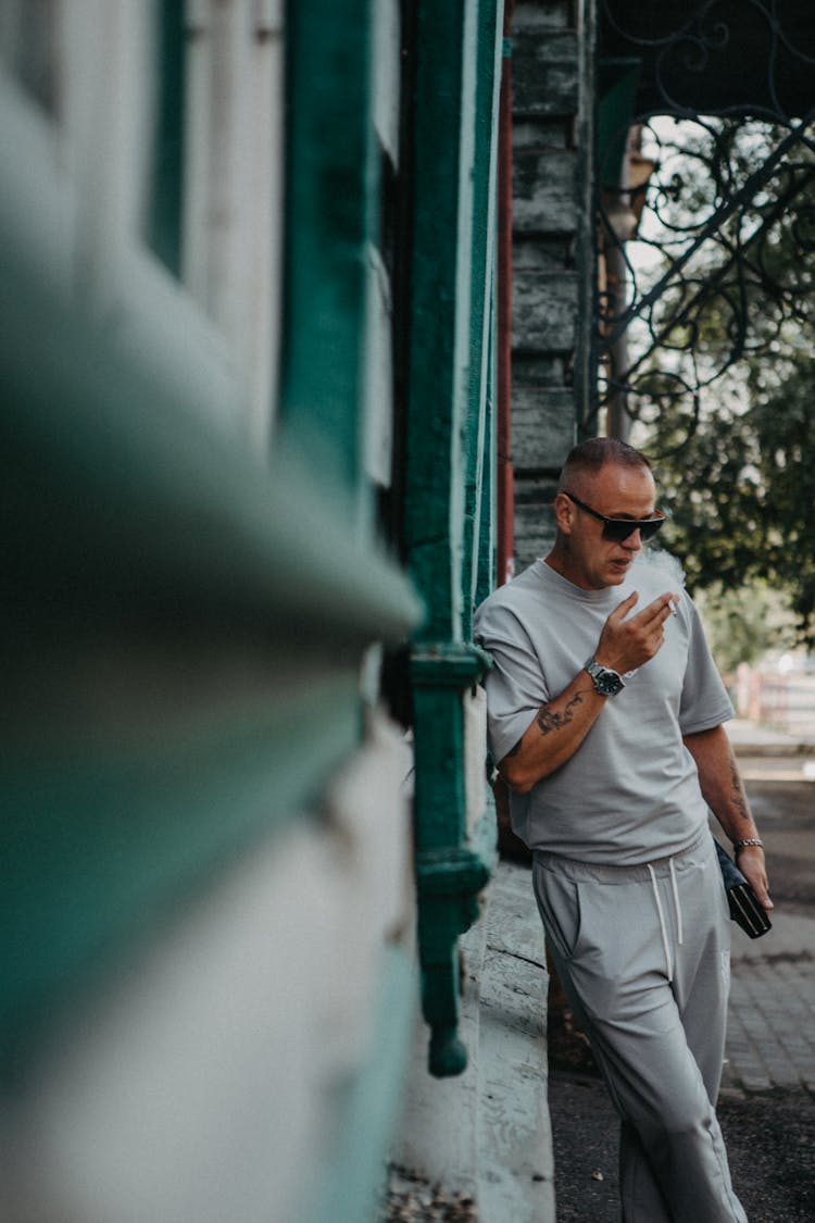 Man In Gray Shirt And Sweatpants Smoking Cigarette While Leaning On The Wall