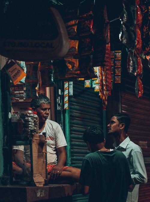 Free Three men engage in conversation at a bustling street kiosk during the night in India. Stock Photo