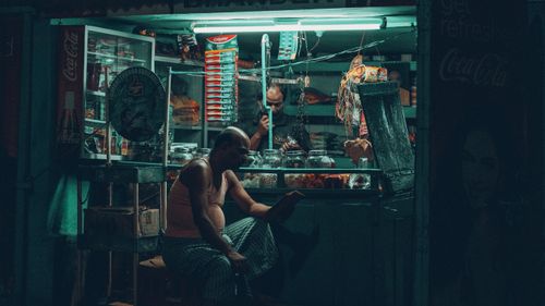 Free A man reading a book outside a vibrant, illuminated small shop at night. Stock Photo