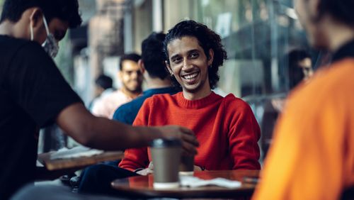 Free A cheerful man in a vibrant red sweater enjoys a coffee with friends at an outdoor cafe. Stock Photo