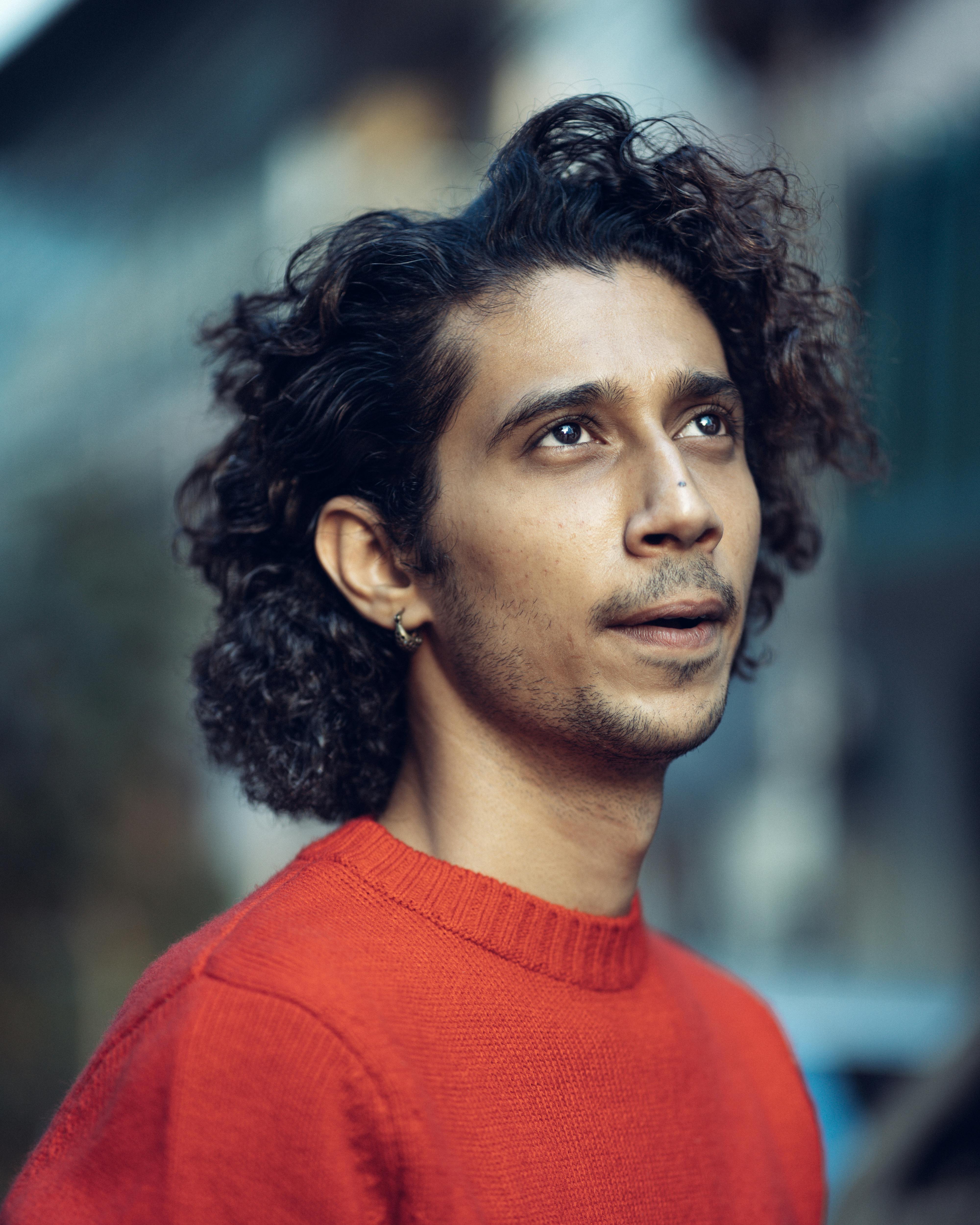 Stylish young man with curly hair in a vibrant red sweater, gazing into the distance outdoors.
