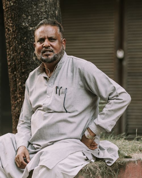 Free Senior man with beard sitting outdoors by a tree, dressed in traditional attire, looking at camera. Stock Photo