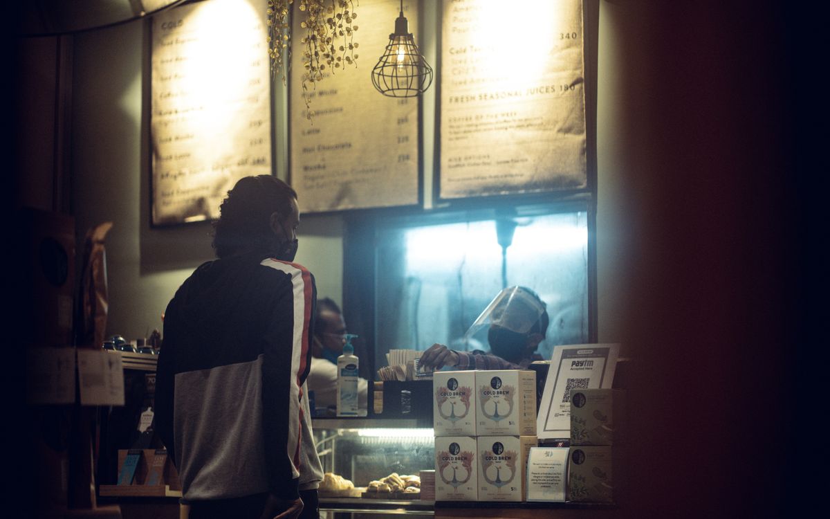 A customer wearing a mask orders at a warmly lit cafe counter during night time.