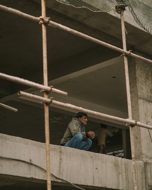 Free Pensive Man in Green Jacket and Blue Denim Jeans Crouching in an Under Construction Building  Stock Photo