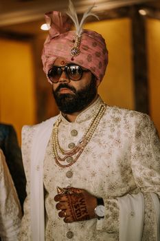 A bearded groom wearing a richly adorned traditional outfit with a pink turban and sunglasses.