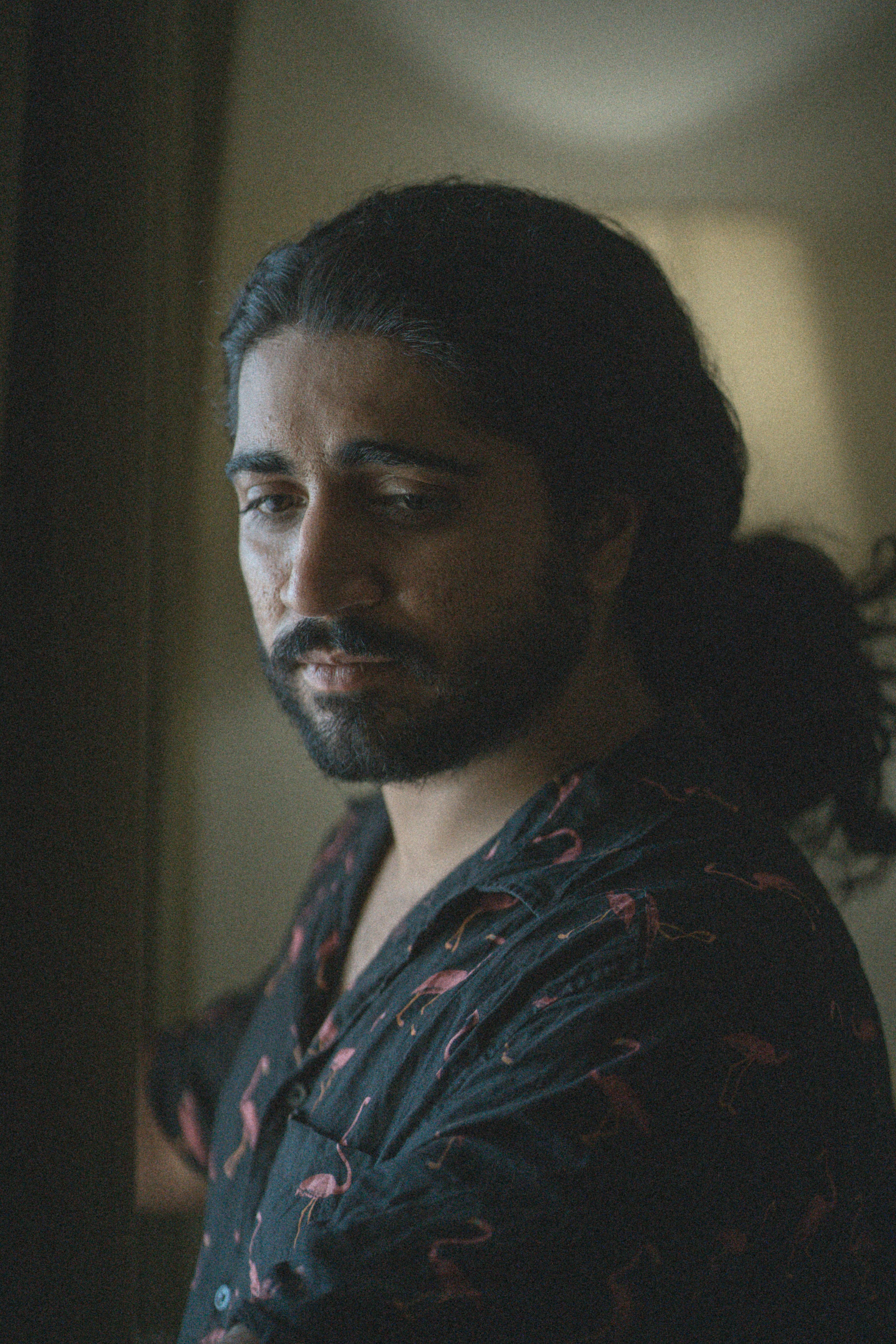 A contemplative man with a beard and long hair gazes thoughtfully out a window indoors.