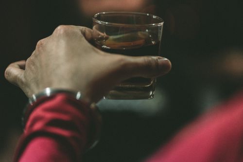 Free A hand in red sleeves holding a glass with a dark beverage, captured in a dimly lit setting. Stock Photo