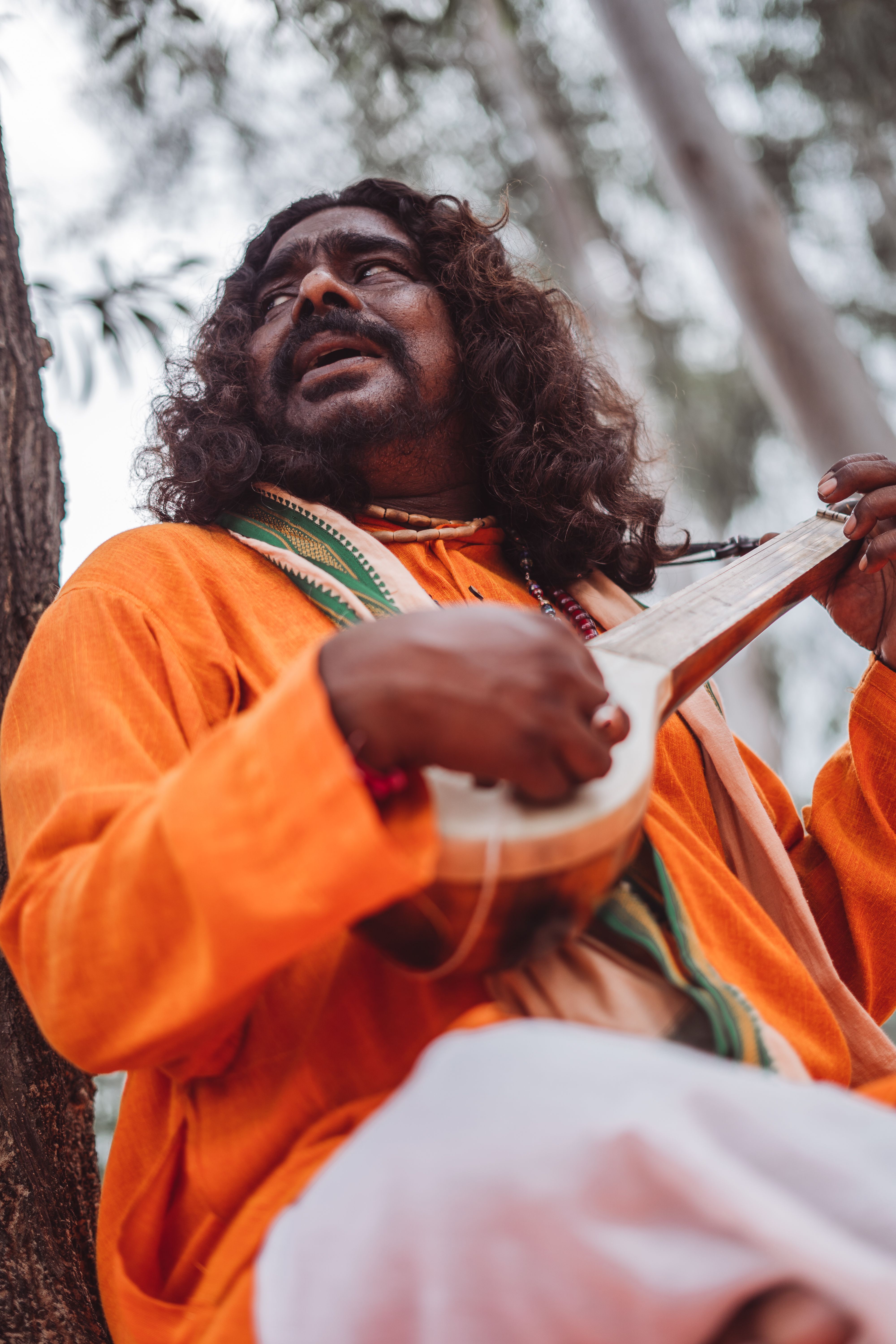 A man in vibrant attire plays a traditional string instrument under a tree, exuding a spiritual essence.