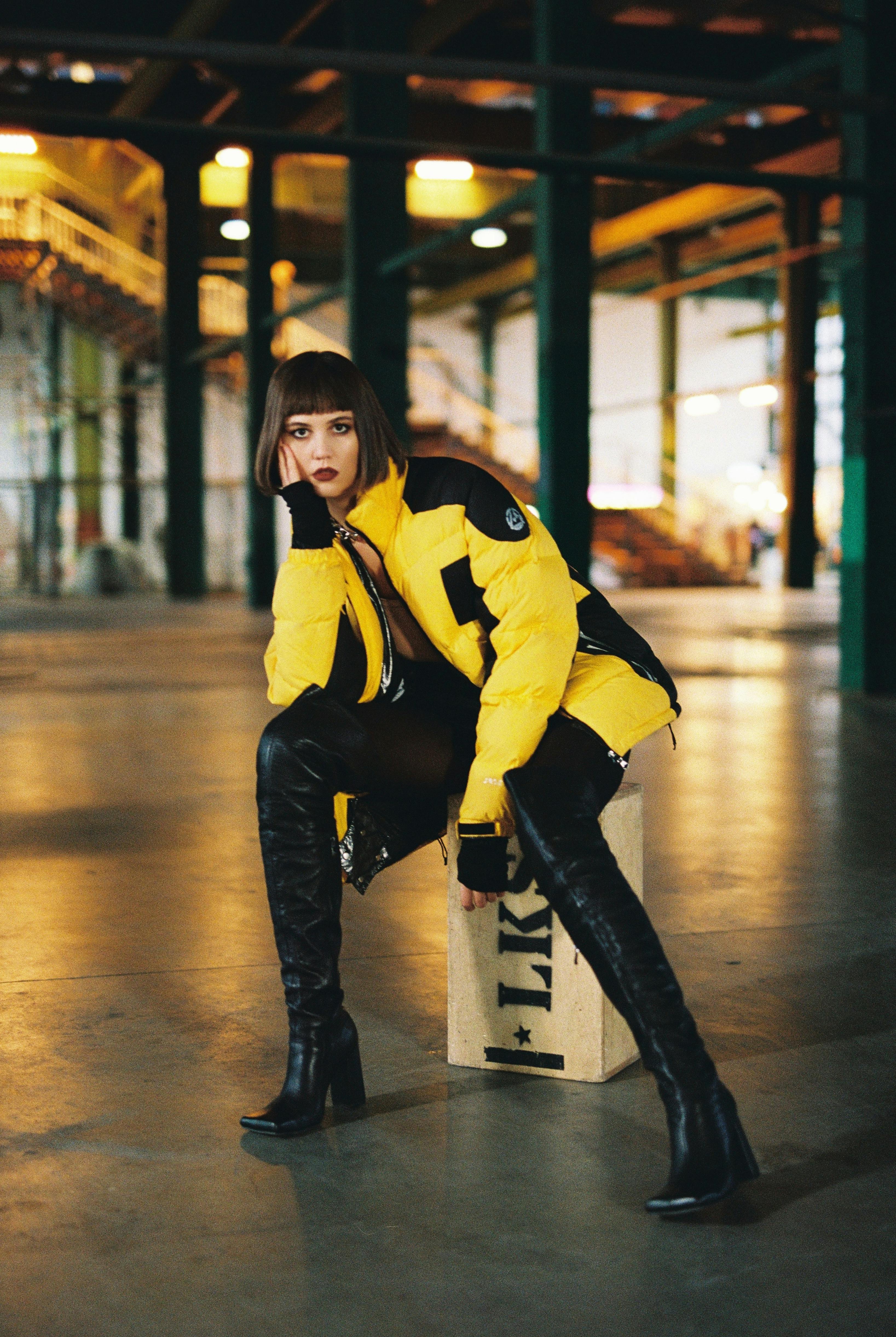 Stylish woman in yellow jacket and boots in an industrial warehouse setting.