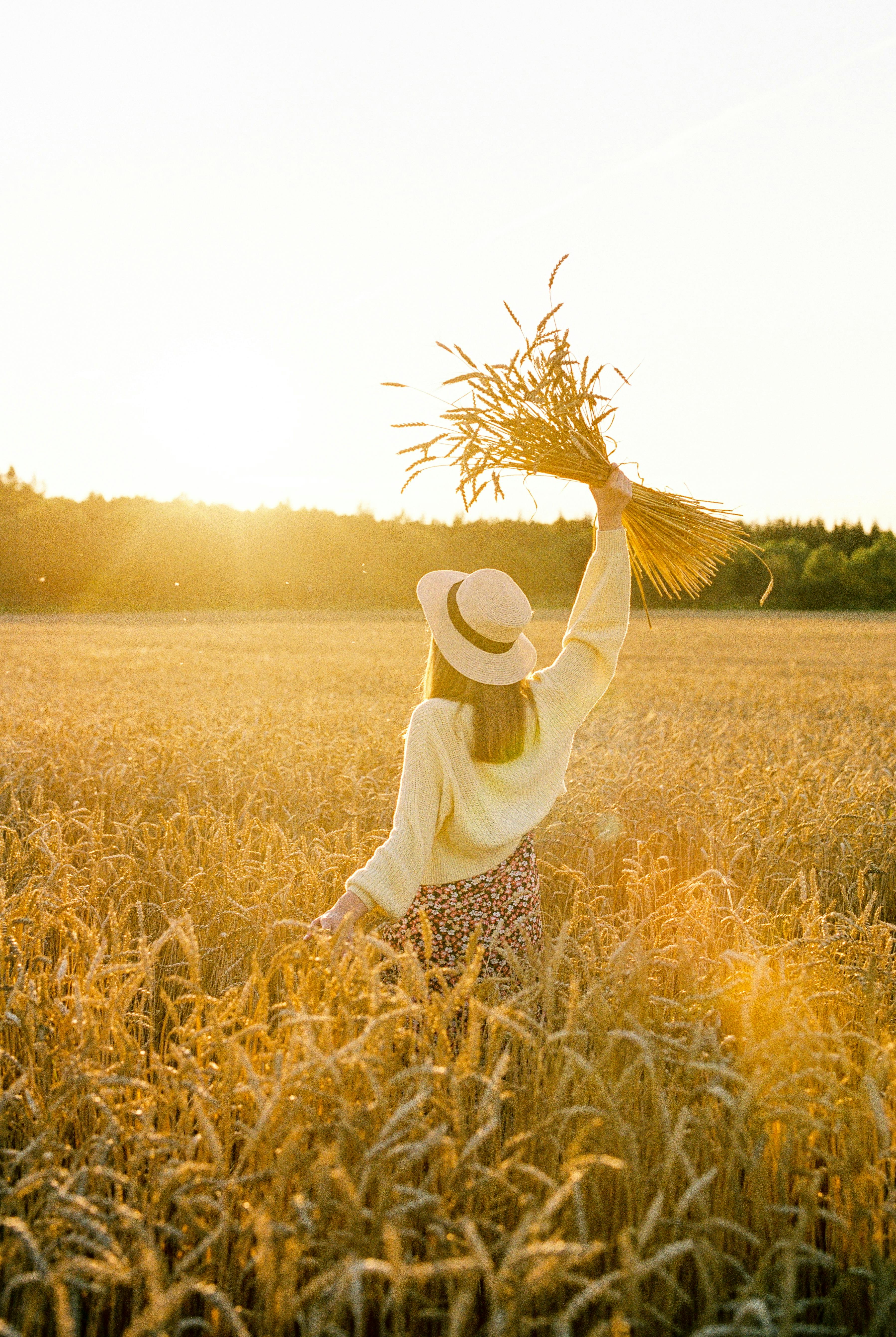 A woman in a hat poses in a wheat field during sunset, celebrating nature's beauty.