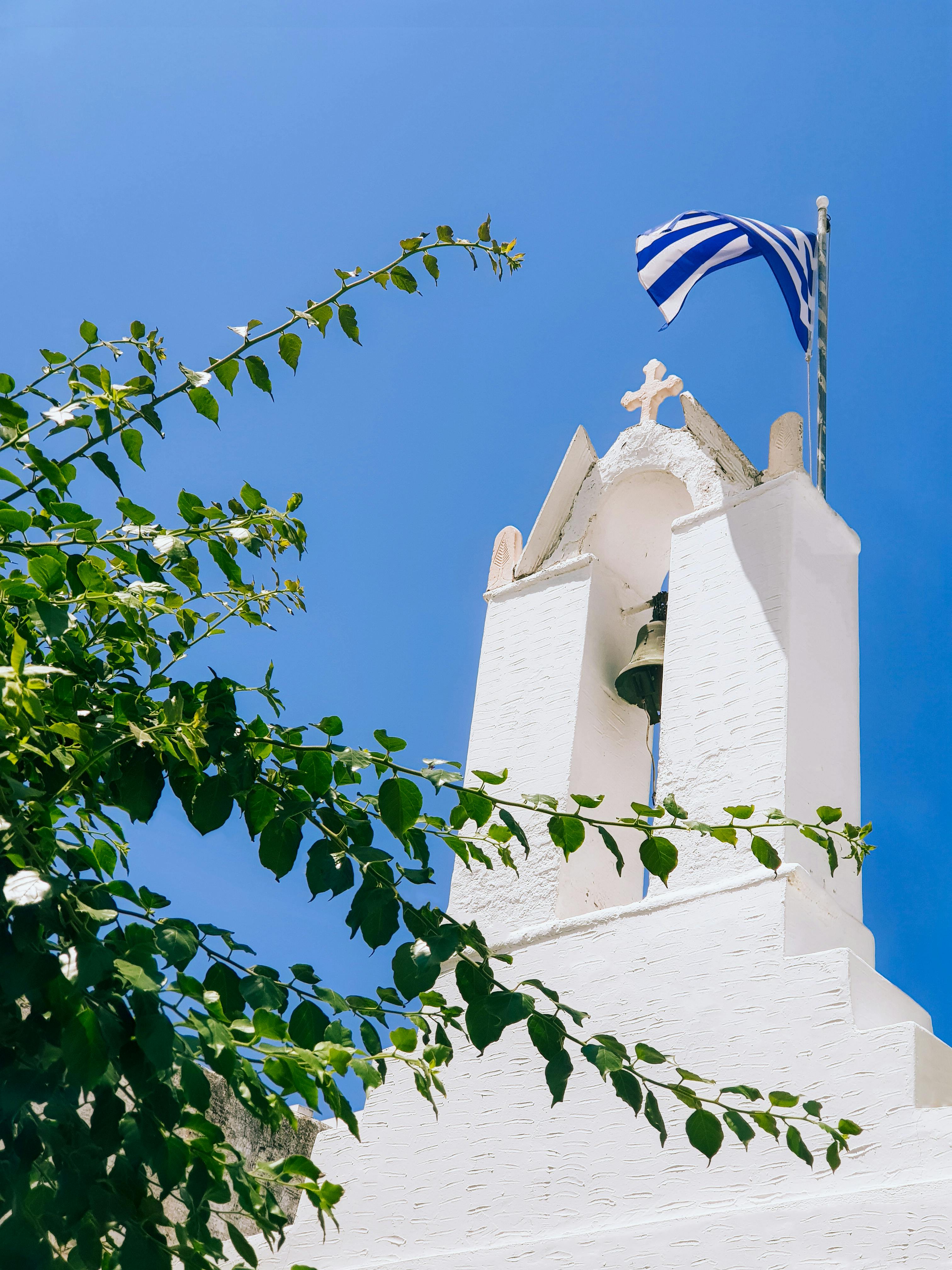 Low-angle view of a white church with a Greek flag in Paros, Greece, under a clear blue sky.
