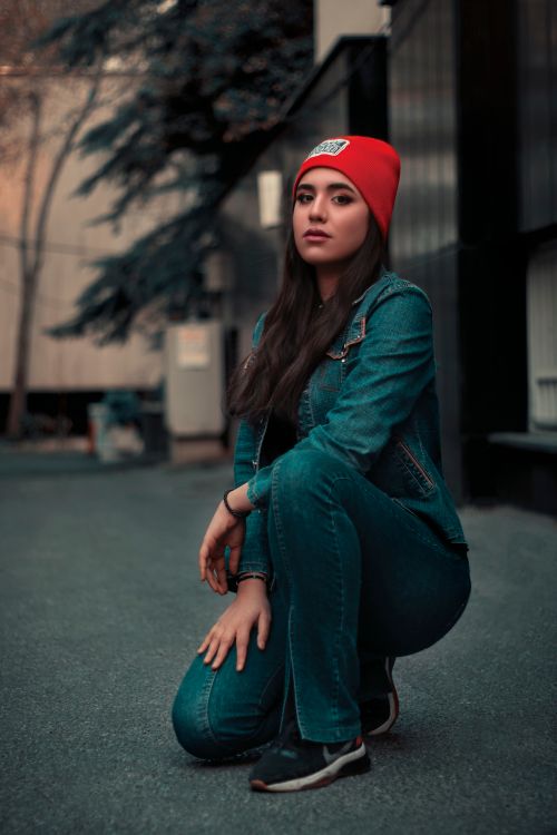 Free Woman kneeling in stylish denim attire with a red beanie, captured in Tehran's urban streets. Stock Photo