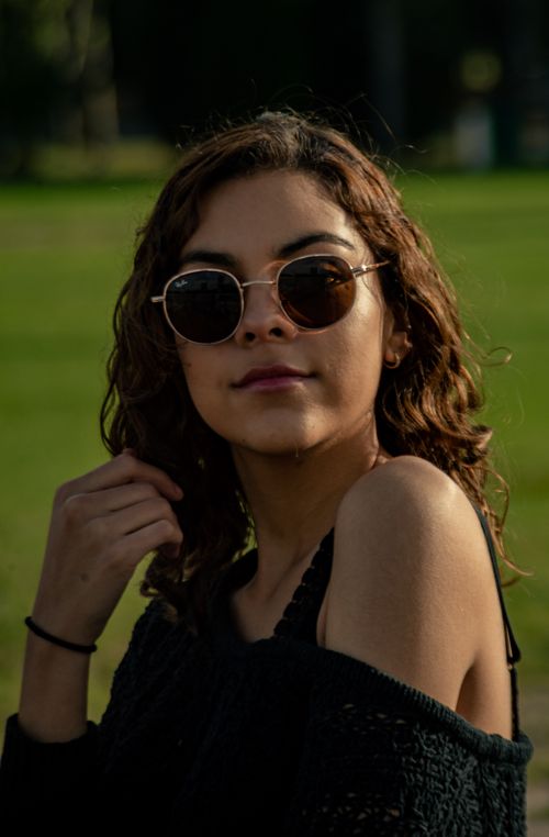 Free A fashionable woman with curly hair poses stylishly in a sunlit field, wearing sunglasses. Stock Photo